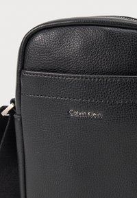 Borsa a tracolla nera in pelle con finitura testurizzata, caratterizzata da un logo "Calvin Klein" in argento e un bordo superiore liscio. Inclusa una tracolla regolabile.
