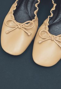 Ballet flats yang terbuat dari kulit cokelat dengan ujung bulat, tepi berenda, dan aksen pita di bagian depan. Lapisan dalam berwarna hitam.