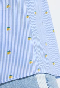 Tissu rayé bleu et blanc avec un petit motif de fruits jaunes, drapé sur un denim bleu clair avec un détail de rayures verticales blanches.