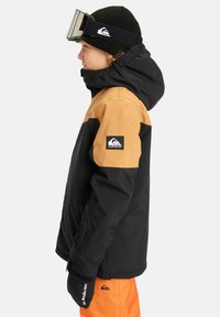 Black and tan snow jacket with a hood, orange pants, and black gloves. Features Quiksilver logo on the arm and ski goggles on the head.
