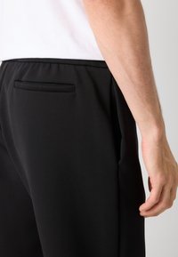 Svarta träningbyxor med en slät yta, elastiskt midjeband och en liten bakficka. Närbild av sidovy och handplacering.