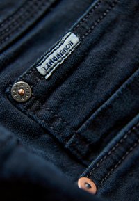 Tummansininen farkkukangas, jossa on teksturoitu pinta, varustettuna brändätyllä tarralla ja kaiverretulla kuparinappulalla. Näkyvät ompeluyksityiskohdat.