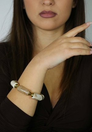 Bransoletka z akcentami w kolorze złotym, ozdobiona białymi i beżowymi koralikami o marmurkowym wzorze oraz wydłużonym, przezroczystym koralikiem centralnym. Noszona na nadgarstku.