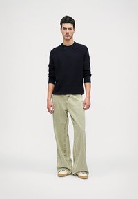 Pulover tricotat bleumarin, pantaloni lejeri de culoare verde deschis și pantofi sport bej. Ansamblul are o croială lejeră și un material texturat.