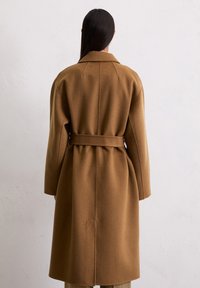 Femme aux longs cheveux noirs portant un manteau en laine camel ceinturé, se tenant face à un mur blanc texturé.