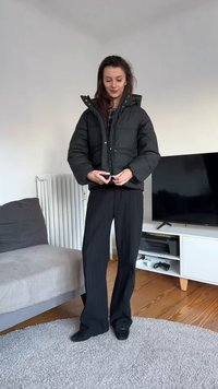 Černá bundička s vysokým límcem a druky, doplněná volnými černými kalhotami. V pozadí je pohovka a televizní stolek.