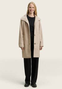 Vrouw die staat en glimlacht, gekleed in een beige knielang jasje over een zwart top en zwarte wijde broek met zwarte schoenen.