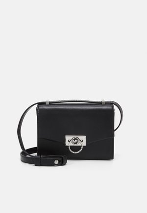 Zwarte leren crossbodytas met zilverkleurige ronde sluiting met MK-logo en verstelbare schouderriem op een witte achtergrond.