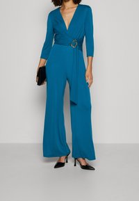 Combinaison bleu canard avec un décolleté en V profond, manches trois-quarts, taille cintrée avec une boucle de ceinture en métal doré et pantalons à jambes larges.