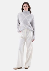 Hellgrauer Strickpullover mit hohem Kragen, der ein Rautenmuster in Creme aufweist. Kombiniert mit weißen weiten Hosen und beigen Schuhen.
