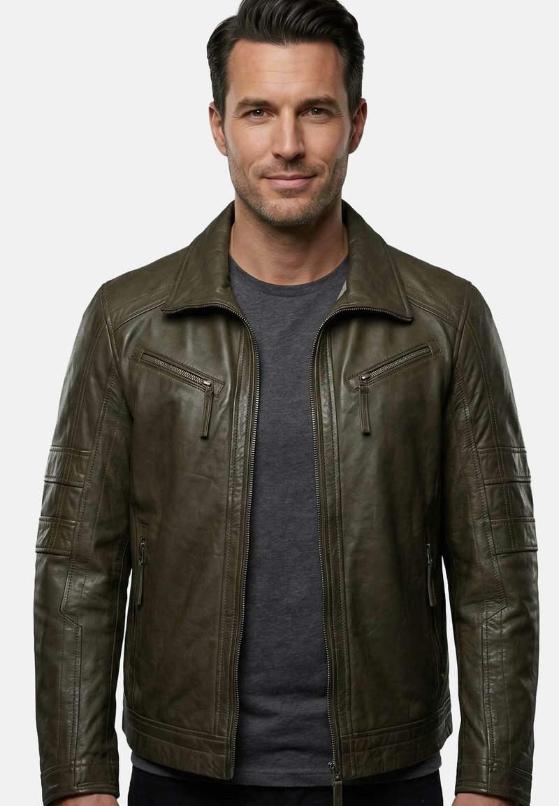 Man with short dark hair wears olive green leather jacket over dark gray shirt, standing against plain light background.