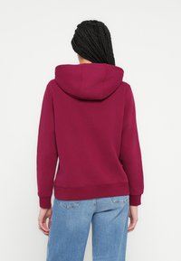 Μπορντό φούτερ με μεγάλη, ρυθμιζόμενη κουκούλα, ριμπ μανσέτες και τελείωμα, κατασκευασμένο από απαλό ύφασμα. Φορεμένο με άνετο, ανοιχτό μπλε τζιν.