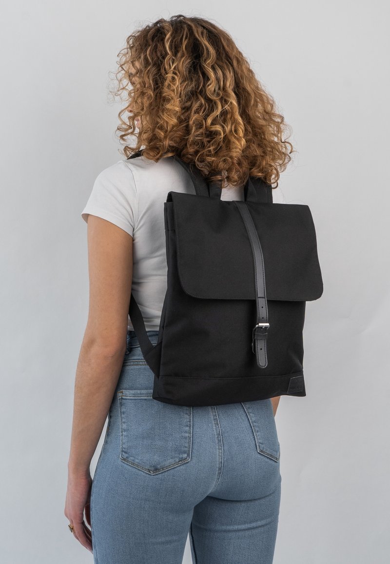 Personne aux cheveux bouclés portant un t-shirt blanc et un jean, avec un sac à dos noir à sangle à boucle sur le dos, sur un fond gris clair.