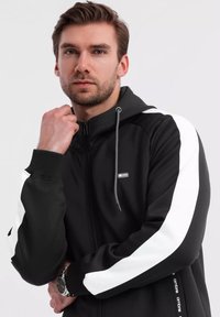 Felpa nera con zip, dotata di strisce laterali bianche, accentuata da un cappuccio con cordino e un patch in gomma con logo. Realizzata in tessuto liscio e testurizzato.