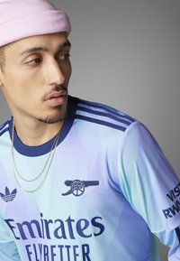 Lys blå fodboldtrøje med mørkeblå accenter og striber, der har "Emirates" logoet og Arsenal kanonemblemet. Guld kædede halskæde synligt.