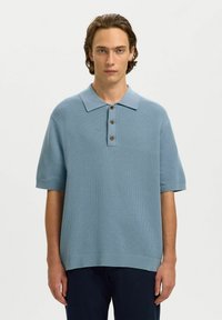 Jeune homme aux cheveux bruns mi-longs portant un polo en tricot à manches courtes bleu clair avec trois boutons et un pantalon foncé.