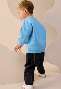 Sweatshirt albastru deschis cu manșete ribbed, asortat cu pantaloni bleumarin cu dungi verticale portocalii și pantofi sport albi. Copil întinzându-se spre o formă.