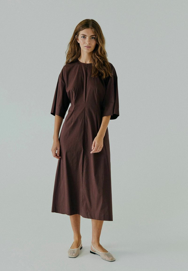 Robe marron, longueur genoux, avec des manches courtes, un col rond et une silhouette droite. Portée avec des chaussures à enfiler beige en tressé.