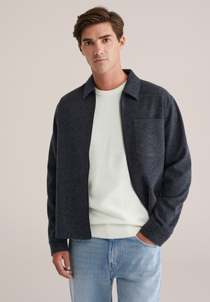 Chaqueta de cremallera de color gris oscuro con cuello tipo solapa, con un bolsillo en el pecho, usada sobre un suéter texturizado de color verde claro, combinada con jeans de color azul claro.