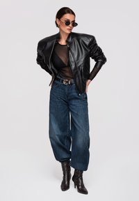 Kobieta ubrana w czarną skórzaną kurtkę, przezroczystą bluzkę, dżinsy z szerokimi nogawkami, czarny pasek, buty na obcasie i okulary przeciwsłoneczne, pozująca na jednolitym tle.