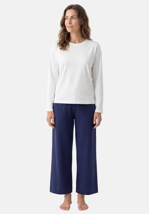 Kobieta o falujących brązowych włosach, ubrana w białą koszulę z długim rękawem i luźne granatowe spodnie, stoi boso na jednolitym tle.