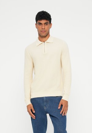 Pullover beige a maniche lunghe con collo zip, texture a coste, colletto piatto e orlo aderente, abbinato a jeans blu.