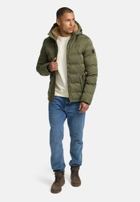 Olive green puffer jacket with a zippered front, fur-lined hood, and side pockets, paired with a cream sweater, blue jeans, and brown boots.