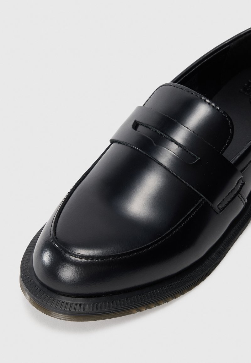 Black leather loafer with a smooth finish, featuring a front strap and stitched accents. The sole has a textured edge for grip.