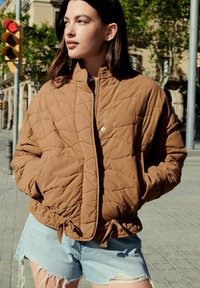 Bruine gestikte pufferjack met een hoge kraag, ritsluiting en trekkoord aan de onderkant. Gecombineerd met lichtblauwe verwerkte denimshorts.