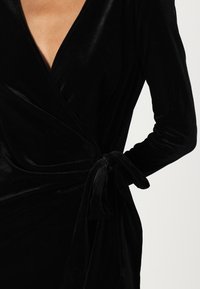 Vestido envolvente de veludo preto com mangas longas, decote em V profundo e detalhe de cintura atada, exibindo uma textura suave e um design fluido.