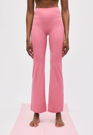 Vrouw staat blootsvoets op een roze yogamat wearing hoge taille, uitlopende roze broek en olijfgroene sportbeha, tegen een effen achtergrond.