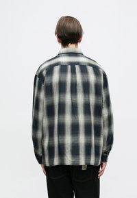 Ilgų rankovių languota marškiniai tamsiai mėlynos ir grietinėlės atspalvių, su laisvu kirpimu, mygtukų apačia ir mažos prekės ženklo etiketės gale.