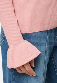 Helles rosa geripptes Sweatshirt mit ausgestellten Ärmeln, kombiniert mit blauen Jeans. Sichtbare Hand, die am Saum des Pullovers ruht.