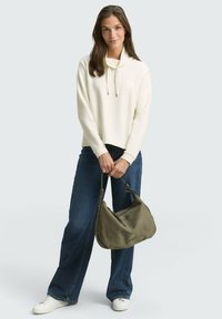 Witte hoodie, relaxte pasvorm, gecombineerd met donkerblauwe wijde jeans. Houdt een olijfgroene tas vast, met witte sneakers aan de voeten.