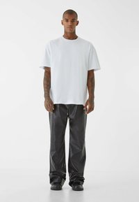 Biela oversized tričko s krátkymi rukávmi, v kombinácii s čiernymi, vysokopásovými, štruktúrovanými nohavicami. Model stojí na bielom pozadí.
