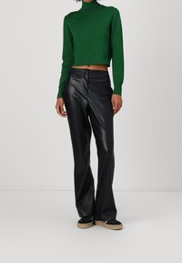 Pull court à col roulé vert en maille douce, associé à un pantalon noir ajusté en simili cuir avec une fermeture éclair frontale et des poignets évasés.