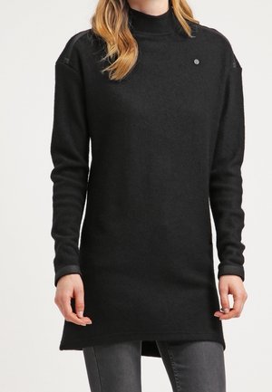Tunic nera a maniche lunghe con collo alto, realizzata in materiale morbido. Presenta un piccolo logo e un orlo leggermente allungato.