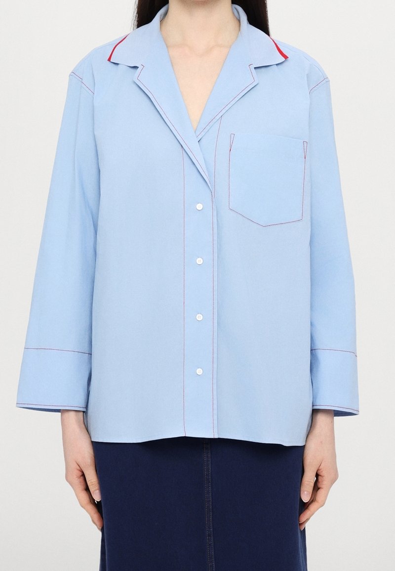 Femme portant une chemise bleu clair à boutons avec coutures rouges et poche poitrine, associée à une jupe bleu marine foncé sur un fond uni.