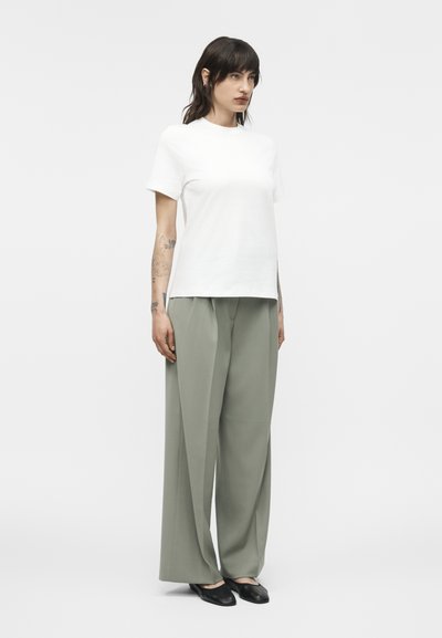 Γυναίκα μοντέλο στέκεται με το πρόσωπο προς τα εμπρός, φορώντας απλό λευκό κοντομάνικο τοπ, φαρδιά ανοιχτογκρι παντελόνια και μαύρα επίπεδα παπούτσια πάνω σε λευκό φόντο.