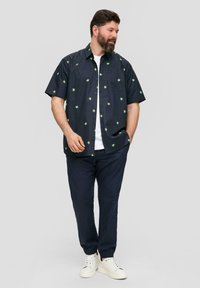 Chemise à manches courtes, de couleur bleu marine foncé avec des motifs de feuilles vertes, devant à bouton, associée à un pantalon marine et des baskets blanches pour un look décontracté.