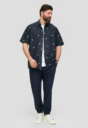 Chemise à manches courtes, de couleur bleu marine foncé avec des motifs de feuilles vertes, devant à bouton, associée à un pantalon marine et des baskets blanches pour un look décontracté.