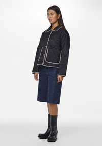 Giubbotto trapuntato nero con piping bianco, bottoni frontali e grandi tasche, indossato con pantaloni corti in denim e stivaletti neri. Texture liscia.