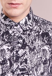Black and white button-up shirt featuring a busy abstract design with various skeletal and organic patterns. Smooth fabric, pointed collar.