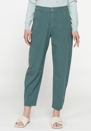 Donna che indossa pantaloni aderenti turchesi, maglia leggera a maniche lunghe e sandali bianchi intrecciati senza lacci, in piedi davanti a uno sfondo semplice.