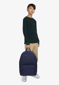 Batoh z tmavě modré látky s přední kapsou a malým červeným logem. Model má na sobě zelené pruhované tričko s dlouhým rukávem a khaki kalhoty.