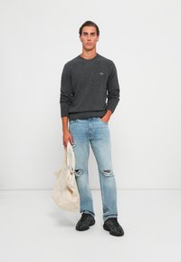 Grå stickad tröja, ljusblå rivna jeans och svarta skor. Modellen håller en beige tote väska. Enkel design utan mönster eller detaljer.