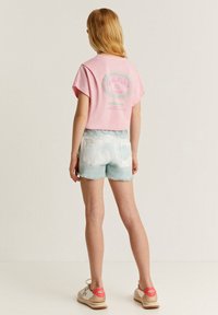 Camiseta corta de algodón rosa con estampado gráfico en la parte trasera, combinada con pantalones cortos desgastados de color azul claro y zapatillas beige con acentos rojos.