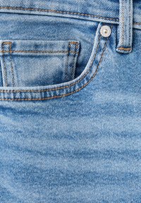Gros plan sur un jean en denim bleu montrant la poche avant, la petite poche à monnaie, la boucle de ceinture et un rivet métallique avec des détails de couture orange.