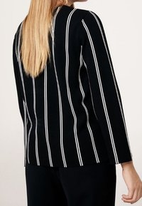 Haut noir à manches longues avec des rayures blanches verticales, présentant une coupe droite et un col rond. Le tissu semble lisse avec une coupe légèrement structurée.