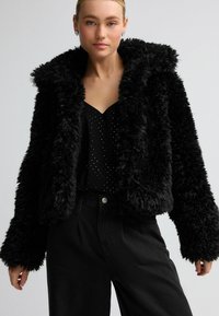Zwarte nepbont cropped jasje met een grote kraag, voorzien van een gestructureerd oppervlak en lange mouwen, gecombineerd met een zwart blouse met stippen en een broek.